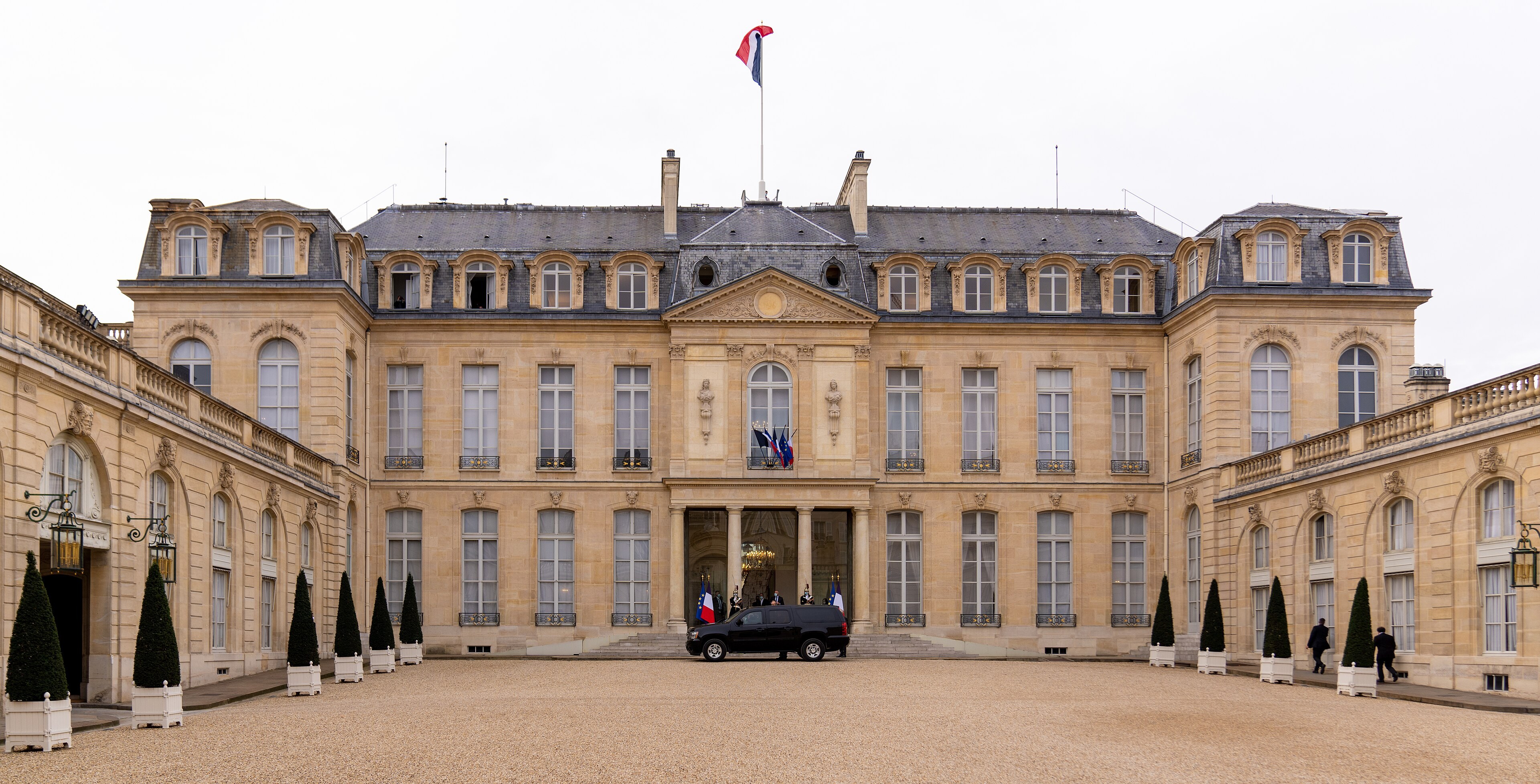 Cour d’honneur de l’Élysée, façade classique, drapeau tricolore, véhicule officiel et deux personnes à droite.