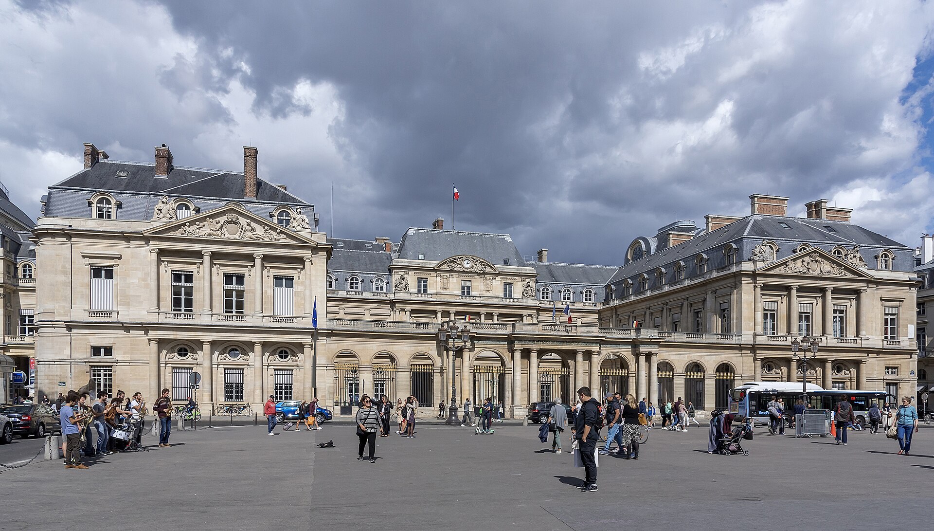 Place du Palais-Royal : ailes du Conseil d’État, grilles dorées, bus et passants sous un ciel chargé.